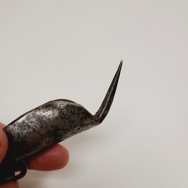 May include: Close-up of a vintage, curved metal tool with a sharp, pointed end. The tool has a worn, aged appearance, with a curved handle and a pointed blade. The metal has a dark, weathered finish, suggesting age and use.