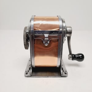 May include: Vintage pencil sharpener with a copper and silver metal finish. The sharpener has a hand crank with a black handle and a gear on the side. The base is rectangular with mounting holes.