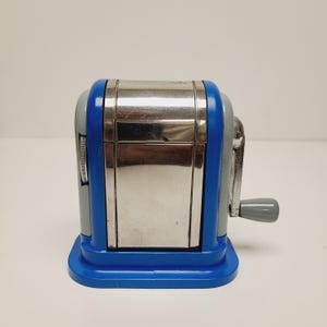 May include: A blue, gray, and silver pencil sharpener with a hand crank. The sharpener has a rectangular shape with a blue base and accents. The crank and other details are silver.