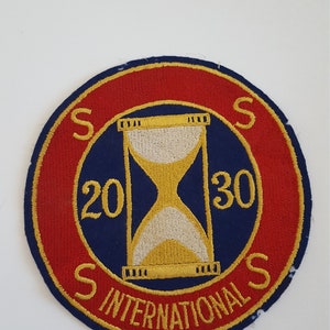 May include: A blue and red circular patch with gold embroidery. The center features a gold hourglass with white sand. The text "SSS 20 30 SSS INTERNATIONAL" is embroidered around the hourglass.
