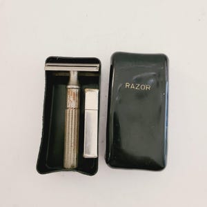 May include: An open black travel case containing a vintage silver razor, a small silver container, and a black case with the word "RAZOR" in gold lettering. The razor has a textured handle.