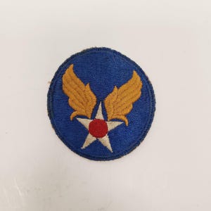 May include: A circular blue patch featuring a white star with a red center and gold wings. The patch is likely a military insignia or emblem, possibly for the Air Force. The patch is embroidered with detailed stitching.