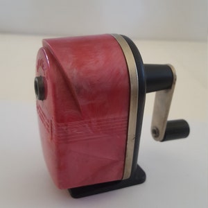 May include: A vintage red pencil sharpener with a black crank and a silver metal band. The sharpener has a textured surface and a small hole for inserting pencils.