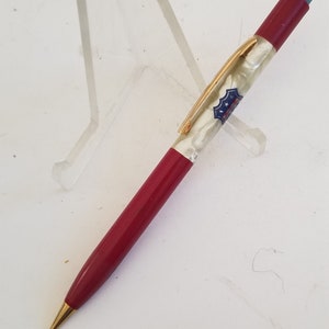 May include: A red and white mechanical pencil with a gold-colored clip and tip. The pencil has a blue eraser and a decorative emblem. The pencil is resting on a clear plastic stand.