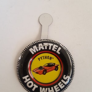 May include: A black and yellow Hot Wheels button with a red car and the words "Mattel", "Python", and "Hot Wheels". The button has a silver ring around the edge and a white tab on the top.