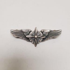 May include: A silver-toned pilot's wing pin with a propeller in the center and detailed wings on either side. The pin is likely made of metal and has a vintage or antique appearance. The design is symmetrical and detailed.