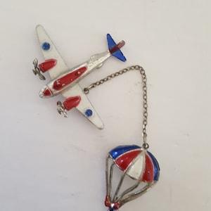 May include: A silver airplane charm with red, white, and blue paint, attached to a silver chain with a red, white, and blue parachute charm.