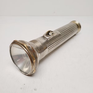 May include: A vintage silver flashlight with a ribbed body and a gold-toned bezel around the lens. The on/off switch is located on the side. The flashlight is designed with a classic, retro aesthetic.