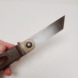 May include: A folding knife featuring a single-edged, flat-tipped metal blade with a visible grain. The handle is constructed from dark brown wood, secured to the blade with a circular brass pivot pin and a small brass thumb stud for opening.