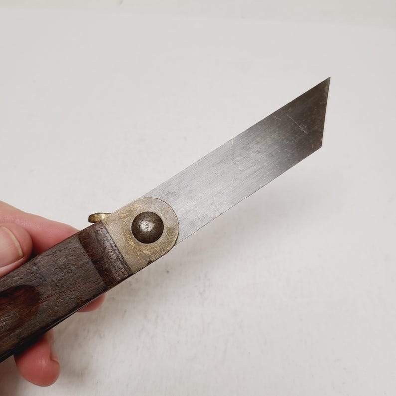 May include: A folding knife featuring a single-edged, flat-tipped metal blade with a visible grain. The handle is constructed from dark brown wood, secured to the blade with a circular brass pivot pin and a small brass thumb stud for opening.