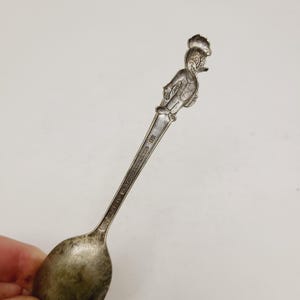 May include: A vintage silver spoon with a cartoon character figure at the top. The spoon has a rounded bowl and a long handle with engraved text. The cartoon figure is smiling and has a hat. The spoon is held in a hand.