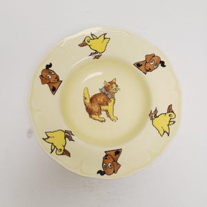 May include: A round, yellow plate with a scalloped edge. The plate features illustrations of a brown and yellow cat, yellow ducks, and brown dogs. The illustrations are in a cartoon style. The plate is likely for children.