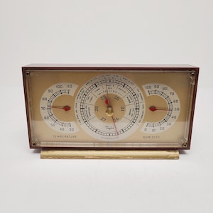 May include: A rectangular Taylor Stormguide weather station with a maroon frame and a gold-toned base. The device features three circular gauges displaying temperature, barometric pressure, and humidity with red needles on a beige background.