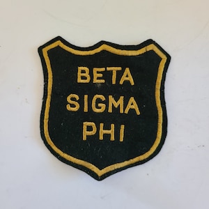 Puede incluir: Un parche negro en forma de escudo con ribete y letras bordadas en oro. El texto "BETA SIGMA PHI" está bordado con hilo dorado. El parche es probablemente para un club u organización.