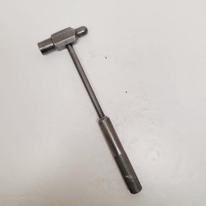 May include: A small, vintage metal hammer with a textured handle. The hammer has a cylindrical head on one side and a rounded head on the other. The handle is long and slender, with a textured grip for secure handling.