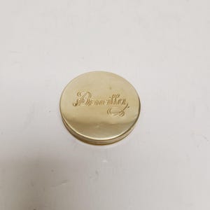 May include: A gold-colored, round lid with the word "Boneilla" elegantly engraved in a cursive font. The lid has a shiny, reflective surface and a simple, classic design. The lid is likely from a vintage cosmetic or beauty product.