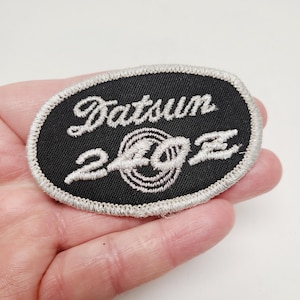 May include: Oval-shaped black patch with the text "Datsun 240Z" embroidered in white thread. The patch has a silver border and features a stylized "240Z" design in the center.