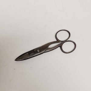 May include: Vintage metal scissors with a dark, aged appearance. The scissors have two finger holes and a pointed blade. The metal has a worn, textured look, suggesting age and use. The scissors are likely for fabric or paper.