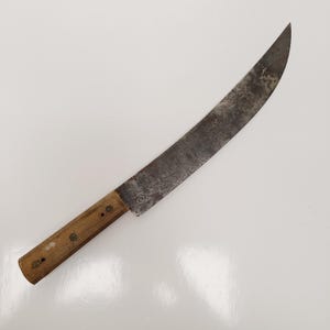 May include: A large, vintage knife with a curved, aged steel blade and a wooden handle. The blade shows signs of wear and rust, indicating its age and use. The handle is attached with small metal rivets.