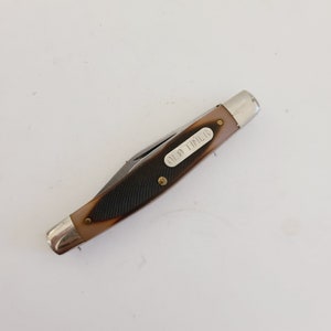 May include: A vintage pocket knife with a brown and black handle. The knife has a silver blade and end caps. The text "OLD TIMER" is printed on the handle. The knife is closed and resting on a white surface.