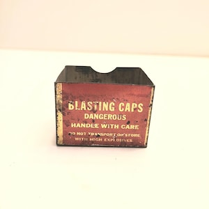 May include: A red metal box with the words "BLASTING CAPS DANGEROUS HANDLE WITH CARE DO NOT TRANSPORT OR STORE WITH HIGH EXPLOSIVES" printed on it.