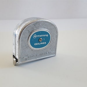 May include: A vintage silver Armstrong tape measure with a blue and white label that reads "Armstrong CEILINGS". The tape measure is marked "ADD 2 INCHES".