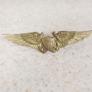 May include: A vintage brass-colored US Navy aviation wings pin. The pin features a shield with vertical stripes, anchors, and wings. The wings are detailed with feather patterns. The pin is likely made of brass or a similar metal.
