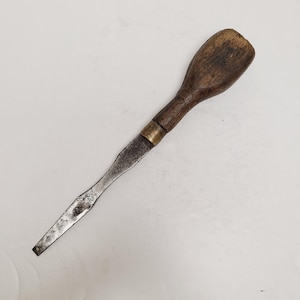 May include: A vintage chisel with a wooden handle and a metal blade. The handle is a dark brown color, and the blade shows signs of wear. A brass band connects the handle to the blade. This tool is likely used for woodworking or carpentry.