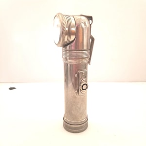 May include: A silver flashlight with a  90-degree angled head. The flashlight has a textured surface and a black switch.