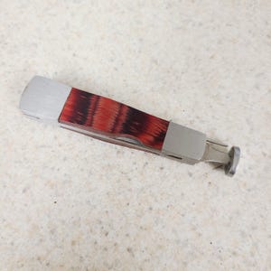 May include: A multi-tool pipe tool with a stainless steel body and a handle made of red and brown wood. The tool has a tamper, reamer, and pick. The tool is approximately 4 inches long.