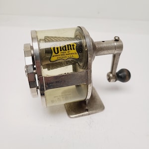 May include: A vintage metal pencil sharpener with a clear plastic casing and a hand crank. The yellow label reads Giant Made By Automatic Pencil Sharpener Co. Chicago, U.S.A. It features a silver metallic finish, a black handle knob, and internal metal blades.
