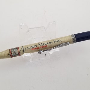 May include: A vintage pencil with the text "ALLIED MILLS, INC. WAYNE FEEDS" printed on the cream-colored body. The pencil has a silver tip, a dark blue end, and a red eraser. A silver clip is attached to the pencil.