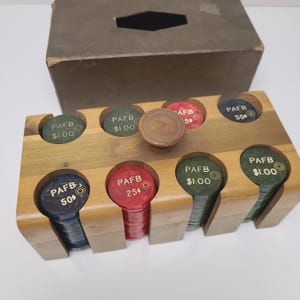May include: A wooden poker chip holder with a variety of chips in red, green, and blue. The chips are labeled with "PAFB" and various denominations. A wooden knob sits in the center. A brown box is in the background.