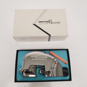 May include: A Dexter Mat Cutter set in its original box. The box is off-white with black text and a graphic. The mat cutter is chrome with a black handle and a red and orange blade. The set is in a black box with a teal interior.