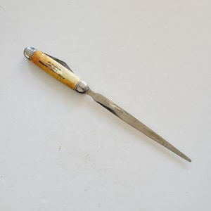 May include: A vintage letter opener with a yellow and white handle. The handle has the text "All Mankind the Bird" and "Mahoney + Cherry + Walker".