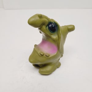 May include: A green, cartoonish figurine of a creature with a large mouth and a pink interior. The figurine has a bulbous head, large eye, and a small arm-like protrusion on its side. The overall design is whimsical and playful.