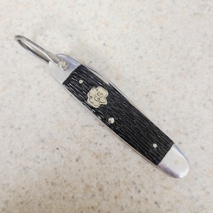 May include: A small, vintage pocket knife with a black textured handle and a silver-colored metal frame. The knife features a loop at one end and a decorative emblem with the letters "GS" on the handle. The blade is folded inside.