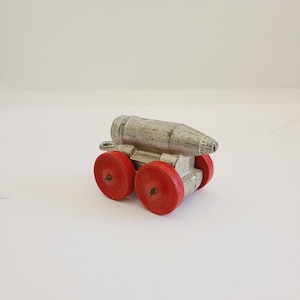 May include: A silver toy cannon with red wheels. The cannon is shaped like a tube and has a small opening at the front. The wheels are made of plastic and have a red rim.