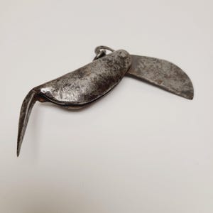 May include: An antique, weathered metal folding knife with a curved blade and a pointed hook. The knife is open, revealing the blade and hook, and has a small ring at the top for hanging. The metal has a dark, aged appearance.