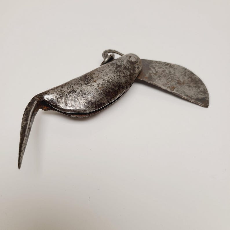 May include: An antique, weathered metal folding knife with a curved blade and a pointed hook. The knife is open, revealing the blade and hook, and has a small ring at the top for hanging. The metal has a dark, aged appearance.