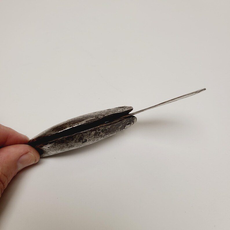 May include: A vintage, weathered metal tool with a long, thin blade extending from a dark, oval-shaped body. The tool appears to be made of iron or steel, with signs of age and use.