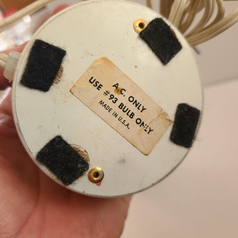 May include: Underside of a small, round, off-white lamp base with four black felt pads and gold-colored screws. A sticker reads "A.C. ONLY #93 BULB ONLY MADE IN U.S.A." Beige electrical cord and plug are visible.