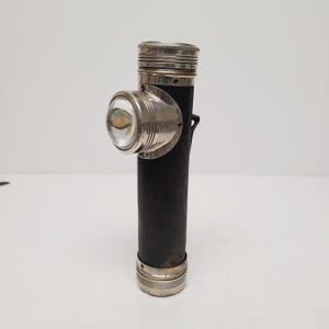 May include: An antique black flashlight with a silver-colored bezel and lens housing. The cylindrical body has a textured pattern. The lens is clear, and the top and bottom of the flashlight are also silver-colored.