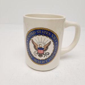 May include: Cream-colored ceramic mug featuring the United States Navy Military Sealift Command emblem. The emblem is a circular design with a blue border, gold rope trim, and a central eagle with a shield. The mug has a curved handle.