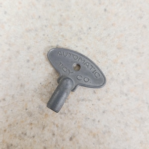 May include: A gray metal wind-up key for a toy, with the text "AUTOMATIC TOY CO." embossed on the head. The key has a small hole and a cylindrical stem. The key is on a light-colored, speckled surface.