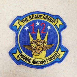 May include: Embroidered patch with a blue background and yellow trim. The patch features the text "THE READY GROUP" and "MARINE AIRCRAFT GROUP". The number "12" is in red, and there is a gold eagle emblem.