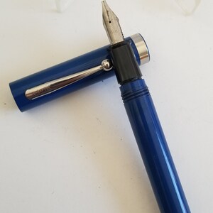 May include: A blue fountain pen with a silver clip and a black nib section. The pen's cap is open, revealing the nib. The pen has the word "SHEAFFER" printed on the clip. The pen is on a white surface.
