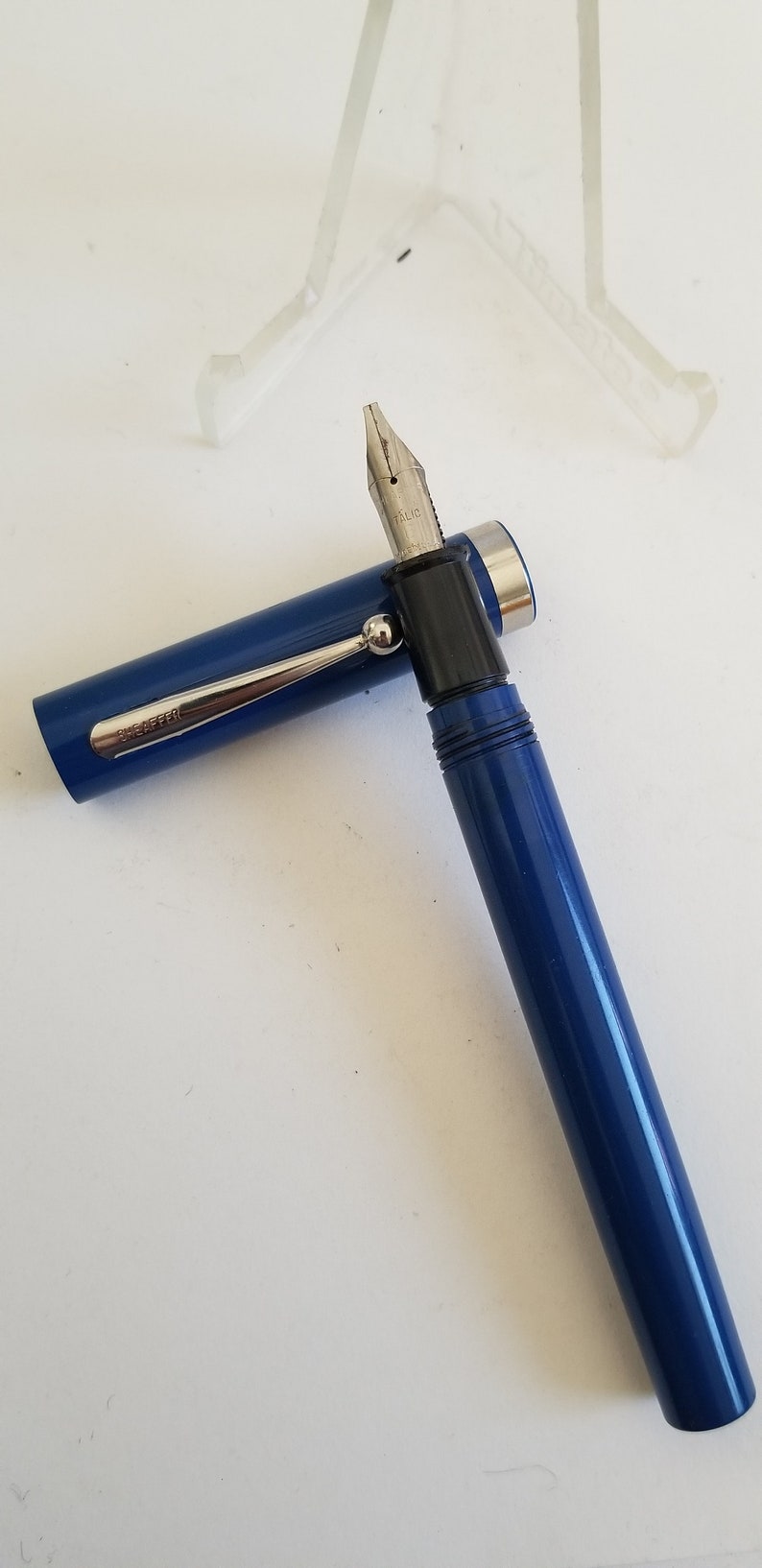 May include: A blue fountain pen with a silver clip and a black nib section. The pen's cap is open, revealing the nib. The pen has the word "SHEAFFER" printed on the clip. The pen is on a white surface.