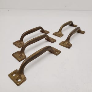 May include: Five antique brass drawer pulls with a curved handle design. Each pull has a rectangular base with two screw holes and a slightly curved handle. The brass has a warm, aged patina.