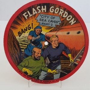 May include: Circular vintage comic book cover featuring Flash Gordon, with the text "City of Sea Caves" and "Part 2." The illustration depicts three characters in a sci-fi setting, with a red spaceship and the word "BANG!"